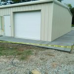 A newly constructed metal garage featuring a white roll-up door and a concrete slab, surrounded by caution tape on the ground.