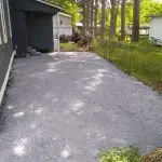 A cleared gravel area beside a house, surrounded by trees and a fence, with an adjoining green lawn.