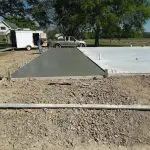 Two construction workers shaping and pouring concrete for a foundation in a rural setting, with vehicles and tools visible in the background.