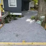 A gravel base is prepared for a pathway outside a house, with garden tools and materials visible nearby.