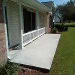 A residential front porch featuring a newly poured concrete slab, brick veneer, and well-maintained landscaping.