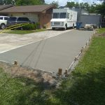 Newly poured concrete driveway with wooden forms and caution tape, surrounded by grass and parked vehicles.