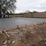 Workers and equipment at a construction site with a freshly poured concrete foundation in the foreground.