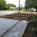 A construction site showcasing an area being prepared for landscaping, featuring wooden frames, soil, and caution tape, with a residential setting in the background.