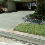 A freshly poured concrete driveway showing wooden edging, with a grassy area alongside and a view of the house in the background.