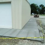 A freshly poured concrete sidewalk is under caution tape beside a metal building, showcasing a clean and smooth surface with an unfinished area in the background.